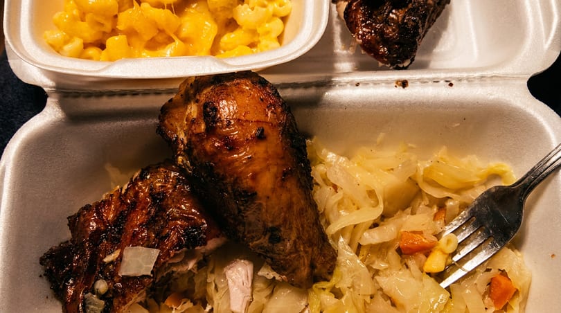 A takeout meal featuring grilled chicken, creamy macaroni and cheese, and stewed cabbage.