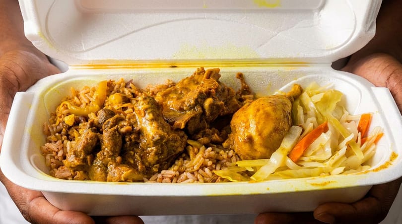 Hands holding a white takeout container filled with curry chicken, rice and peas, and steamed cabbage.