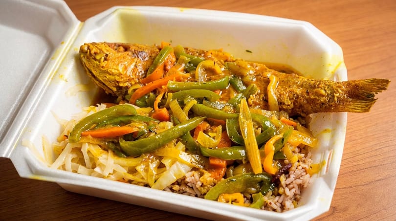 Whole fried fish with colorful peppers, carrots, and onions over rice and cabbage in a takeout container.