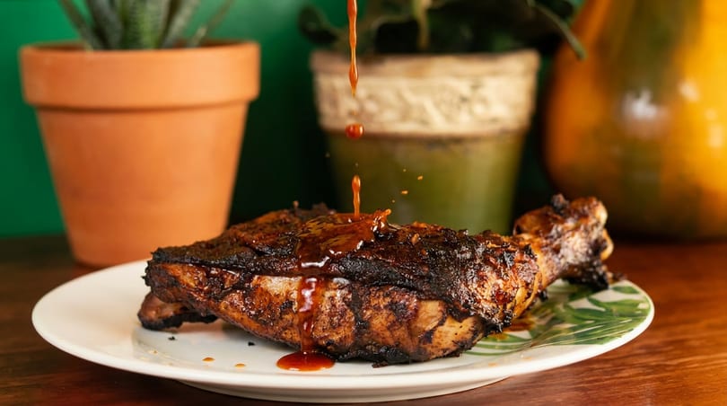 Grilled chicken leg and thigh being drizzled with a rich, dark sauce on a decorative plate.