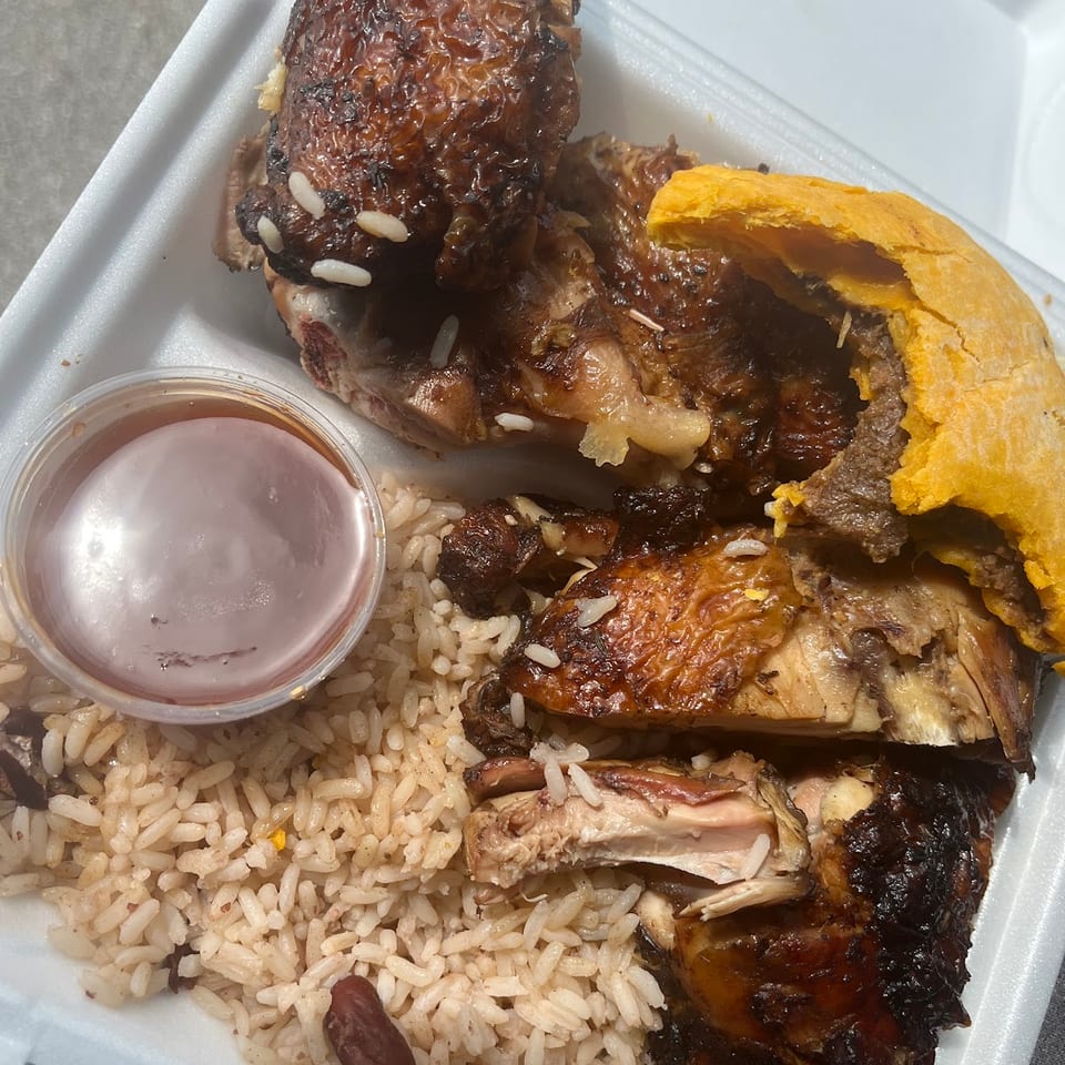 A takeout meal featuring glazed jerk chicken, rice and peas, a beef patty, and sauce.