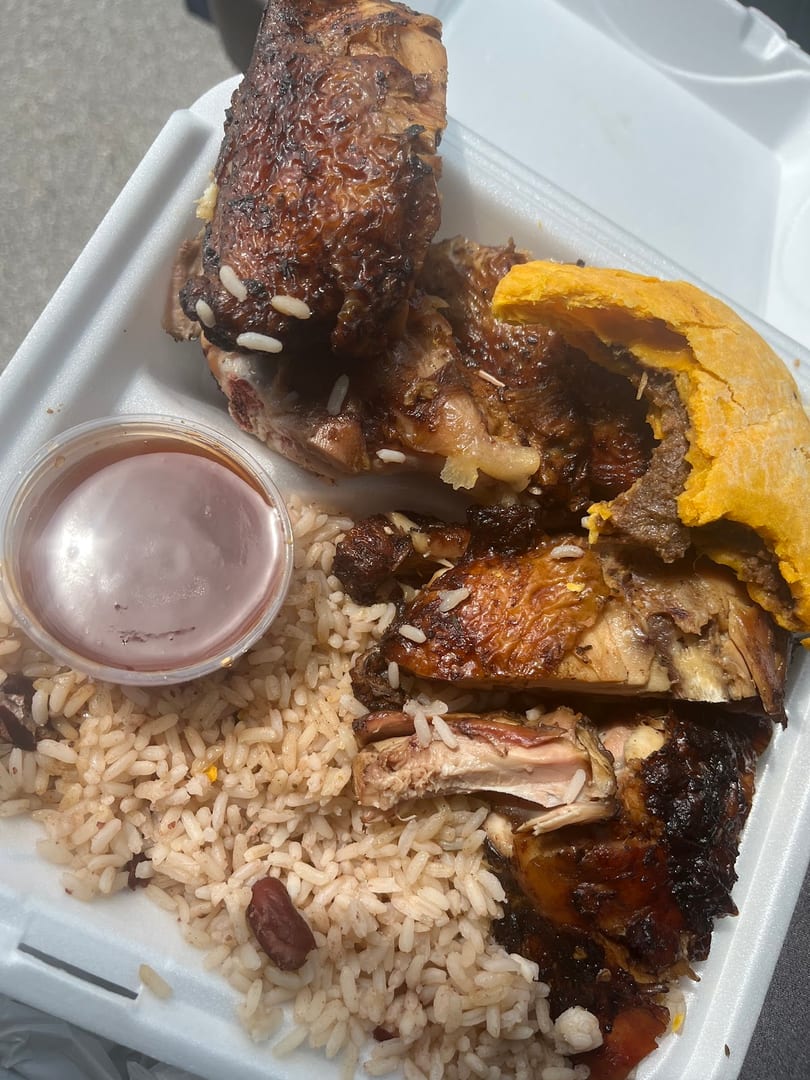 A takeout meal featuring glazed jerk chicken, rice and peas, a beef patty, and sauce.