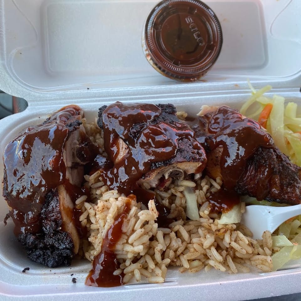 Jerk chicken with sauce, rice, and cabbage in a white takeout container, with an extra sauce cup.