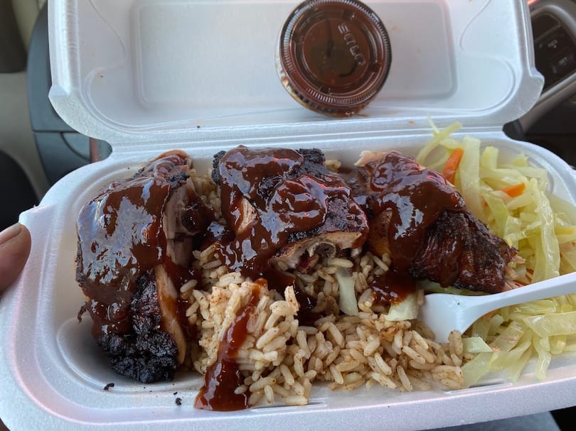 Jerk chicken with sauce, rice, and cabbage in a white takeout container, with an extra sauce cup.